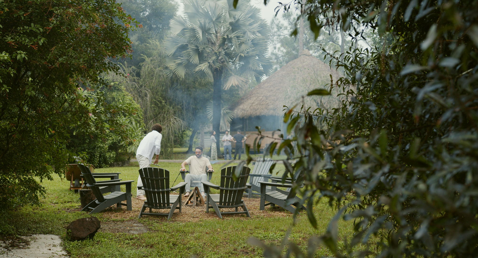 A fire burning in a ring of chairs on the grass at dusk, the maloka behind it through the trees.