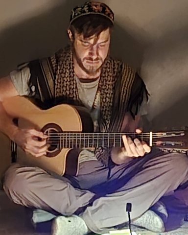 Shlomo Lehrfield seated cross-legged on the floor, playing an acoustic guitar.